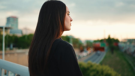 Portrait of cute girl with freckles and loose brown hair wearing black sports hoodie looks at the camera and walks on the bridge of the modern city backgroundの写真素材