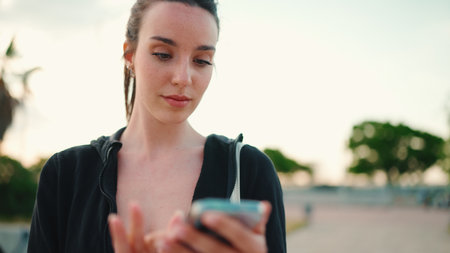 Young woman with freckles and long ponytail wearing black hoodie and beige sports top uses mobile phone and listens to music while standing on bridge modern city backgroundの写真素材