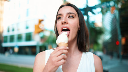 Close-up beautiful woman with freckles and dark loose hair wearing white top testing ice cream. Cute girl enjoy ice cream on modern city backgroundの写真素材