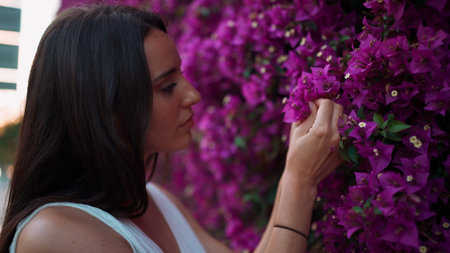 Beautiful woman with freckles and dark loose hair wearing white top walks along wall blooming with...