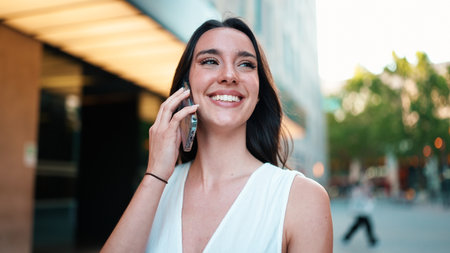 Beautiful woman with freckles and dark loose hair wearing white top is walking down the street with smartphone in her hands. Cute girl talking on mobile phone on modern city backgroundの写真素材