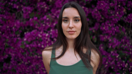 Beautiful woman with freckles and dark loose hair wearing green top poses wall in bloom with purple...