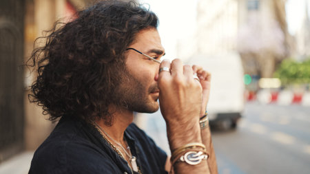 Guy with long curly hair and stubble puts on sunglasses while standing on the sidewalk in front of...