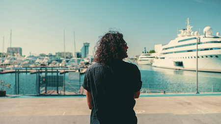 Young italian guy with long curly hair and stubble sits in harbor and looks at ships and yachtsの写真素材