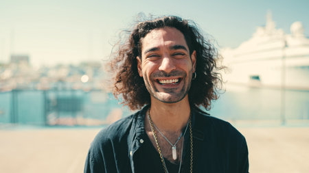 Clouse-up, smiling young italian guy with long curly hair and stubble sits resting on the embankment in the harbor on yachts, ships backgroundの写真素材