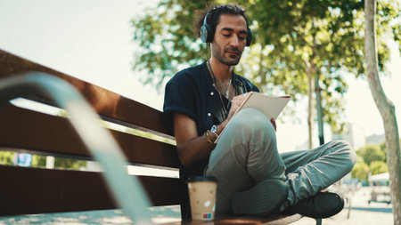Young italian guy with ponytail and stubble sits with headphones on street bench listens to music...