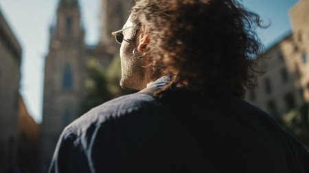 Young italian guy with long curly hair and stubble walks along the fenced sidewalk and looks around.