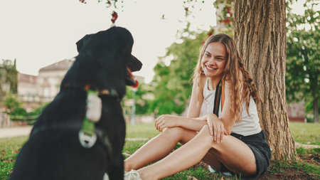 Cute girl with black dog is resting under tree on the lawn. Happy dog rejoices being on walk in the...