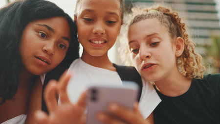 Three girls friends pre-teenage are sitting on the waterfront using mobile phone, watching videos, photos. Three teenagers on the outdoors in urban cityscape backgroundの写真素材