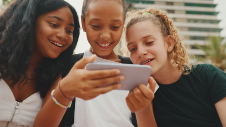 Three girls friends pre-teenage are sitting on the waterfront using mobile phone, watching videos,...