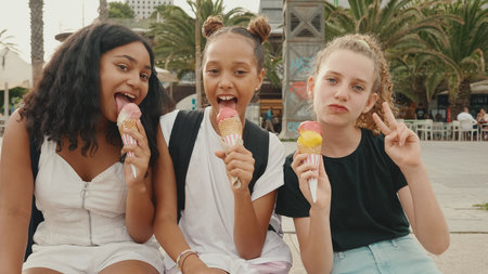Laughing three girls friends pre-teenage are sitting on the waterfront testing ice cream. Three teenagers enjoy ice cream on hot summer dayの写真素材