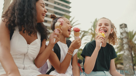 Laughing three girls friends pre-teenage are sitting on the waterfront testing ice cream. Three teenagers enjoy ice cream on hot summer day.の写真素材