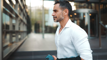 Mature businessman with neat beard wearing white shirt leaves the office in the financial district...