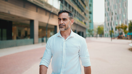 Mature businessman with neat beard wearing white shirt on his way to the office in the financial district in the city. Successful man Looks at the upper floors of modern buildingsの写真素材