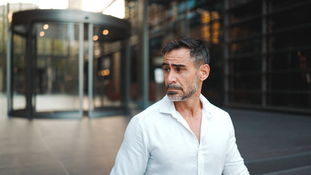 Mature businessman with neat beard wearing white shirt leaves the office in the financial district in the cityの写真素材