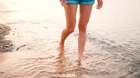 Slim legs of young woman walking barefoot on the beach. Slender female legs and feet walking along the waves of sea water on sandy beachの写真素材