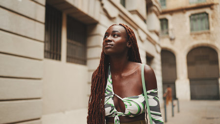 Gorgeous woman with African braids dressed in top walks down the street. Stylish smiling girl looks at the upper floors of buildingsの写真素材