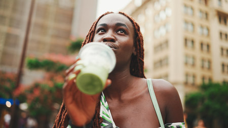 beautiful woman with African braids dress wearing top walks down the street with cold drink in her hands. Stylish girl enjoys drinking fresh cocktail drink in plastic cup with strawの写真素材
