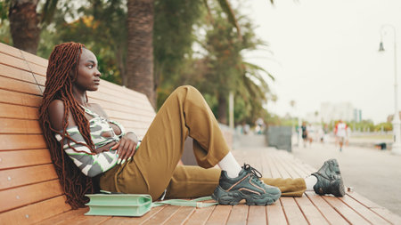 Profile gorgeous woman with African braids wearing in top and trousers is resting sitting on benchの写真素材