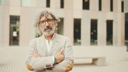 Mature businessman with beard in eyeglasses wearing gray jacket walks down the street past modern buildingsの写真素材