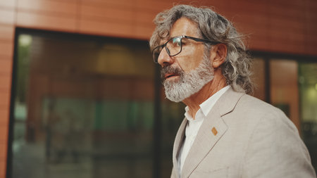Mature businessman with beard in eyeglasses wearing gray jacket walks down the street past modern buildingsの写真素材