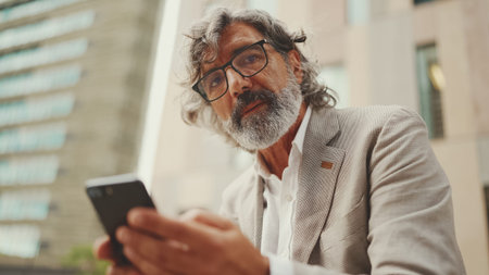Mature businessman with beard in eyeglasses wearing gray jacket holds cellphone in his hand and...