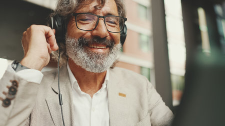 Mature businessman with beard in eyeglasses and headphones, sits in an outdoor cafe and works using tabletの写真素材