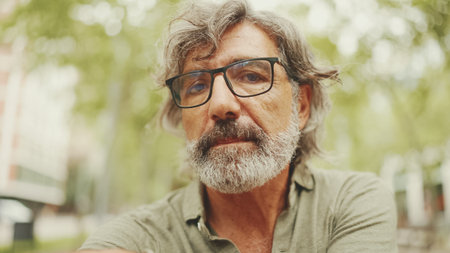 Thoughtful middle-aged man with gray hair and beard wearing casual clothes sits on bench. Mature...