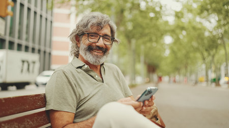Friendly middle-aged man with gray hair and beard wearing casual clothes sits on bench. Mature gentleman in eyeglasses looks at the camera and smilesの写真素材