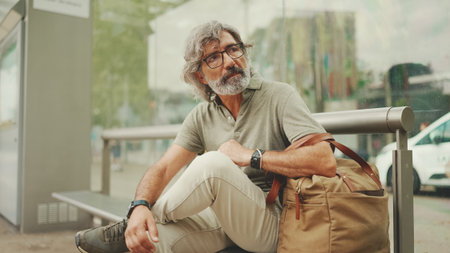 Thoughtful, middle-aged man with gray hair and beard, wearing casual clothes, sits at public...