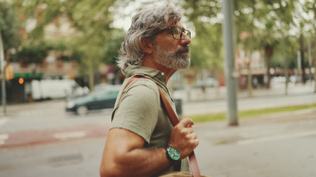 Middle-aged man with gray hair and beard walks and looks around. Mature gentleman in eyeglasses with bag on his shoulder walks through the square on the cityscape backgroundの写真素材