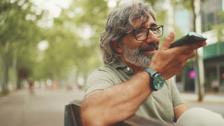 Clouse-up, smiling middle-aged man with gray hair and beard sits on bench and uses mobile phone....