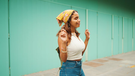Cute tanned woman with long brown hair wearing white top and yellow bandana listens to music in wireless headphones and dances on blue wall background.の写真素材