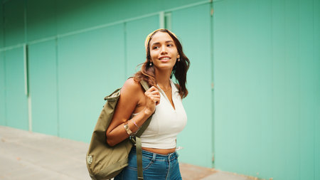 Beautiful girl with tanned skin, brown long hair wearing wireless headphones. Cute young woman with bandana on her head and backpack on her shoulder listens to musicの写真素材