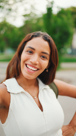 Clouse-up, lovely girl with brown long hair, tanned skin wearing white top is using mobile phone. Smiling young woman taking selfie on cellphoneの写真素材