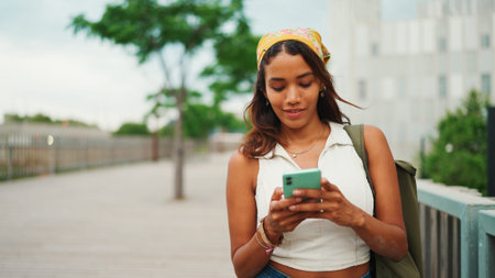 Cute tanned woman with long brown hair wearing white top and yellow bandana walks on bridge with...