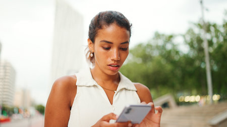 Clouse-up, cute tanned woman with ponytail wearing white top looking up at street signs and map trying to find her way using cellphone. Girl using map application outdoorsの写真素材