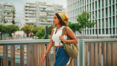Ð¡ute tanned woman with long brown hair wearing white top and yellow bandana walks on bridge with...