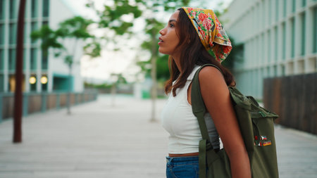 Ð¡ute tanned woman with long brown hair in white top and yellow bandana with backpack on her shoulder and looks around the modern city.の写真素材