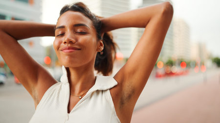 Smiling cute tanned woman with long brown hair wearing white top raises her hands and touches her...