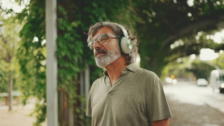 Friendly middle-aged man with gray hair and beard wearing casual clothes listening to music on headphones. Mature gentleman in eyeglasses enjoys music outdoorsの写真素材