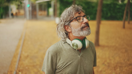 Thoughtful middle-aged man with gray hair and beard wearing casual clothes with headphones around his neck is walking down the streetの写真素材