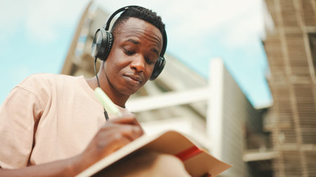 Young african student sitting outside of university and taking notes while studying online with...