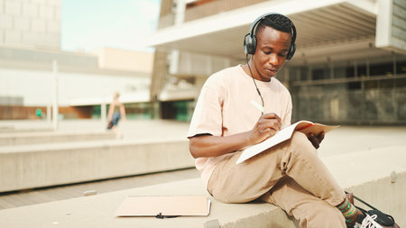 Young african student sitting outside of university and taking notes while studying online with headphonesの写真素材