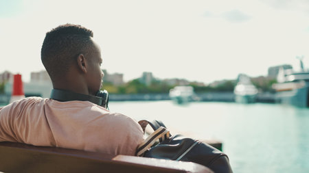 Young african guy relaxes sitting on bench in the port on yachts and ships background. back view
