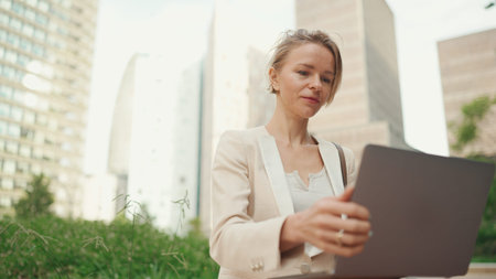 Smiling businesswoman with blond hair wearing beige suit is using pc laptop outside
