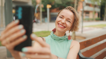 Beautiful happy smiling woman with short blond hair in casual wear sits on bench, makes video call on cellphoneの写真素材