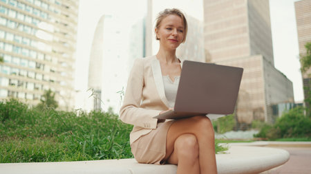 Smiling businesswoman with blond hair wearing beige suit is using pc laptop outsideの写真素材