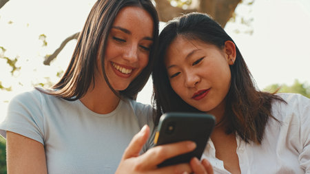 Young multi ethnic smiling women are watching videos on their phone. Girls laugh at picnic on summer day outdoorsの写真素材