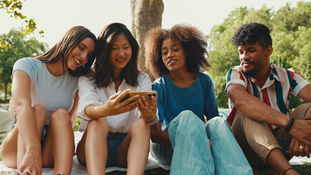 Happy, smiling multiethnic young people at picnic on summer day outdoors. Group of friends talking, using cellphone while relaxing in the park at picnicの写真素材
